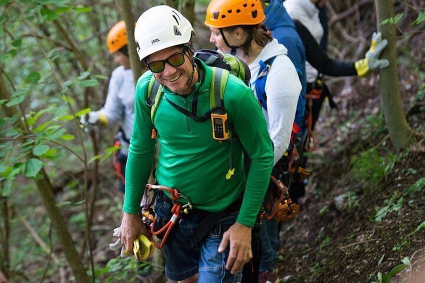Una persona con camisa verde y casco blanco asciende por un sendero boscoso junto a otros escaladores. 2