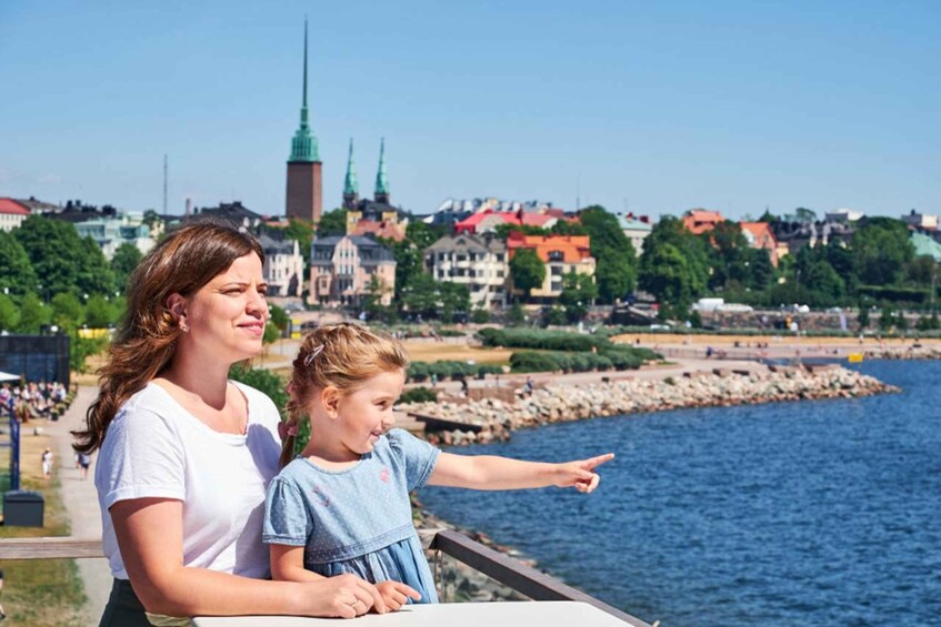 A woman and a child overlooking a waterfront with buildings and a tower in the background. 1