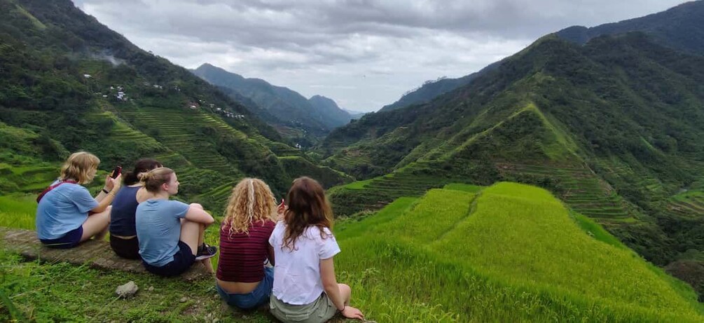 A group of people sitting on a grassy hillside overlooking terraced fields and mountains. 4