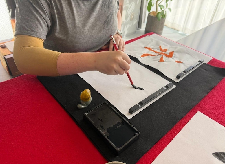 A person painting on a white canvas with a red brush, a small sculpture, and a black tray with paint on a red and black table. 2