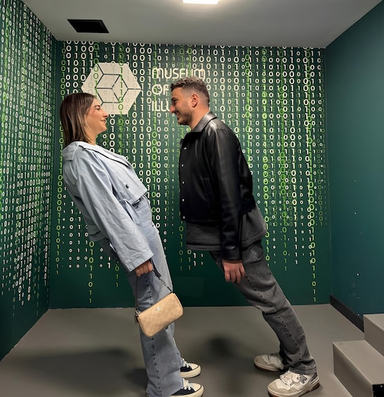 Two people in a room with a green wall covered in binary code patterns. 5