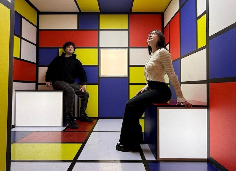 Two people sitting on white blocks in a room with colorful geometric patterns on the walls and floor. 1