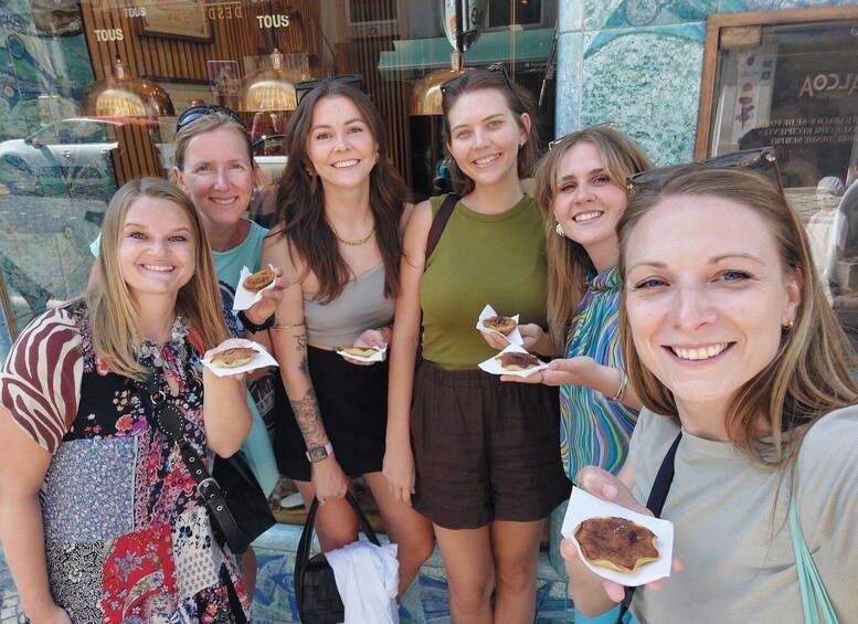 A group of people holding pastries in front of a shop with a sign that reads "TOUS". 1