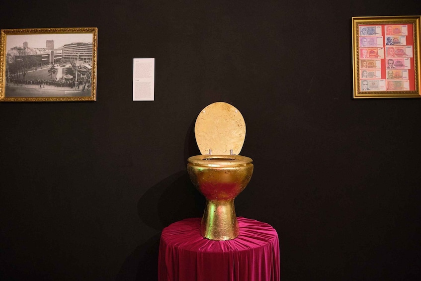 A golden toilet on a red cloth, surrounded by framed pictures and a document on a dark wall. 4