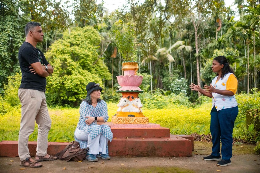 Three people standing and sitting near a temple with a lotus pedestal in a lush garden. 4