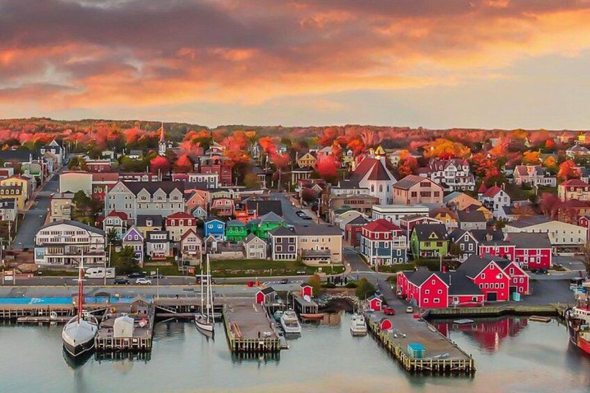 Eine Stadt am Wasser mit bunten Häusern, einem Steg mit Booten und Herbstbäumen im Hintergrund. 2