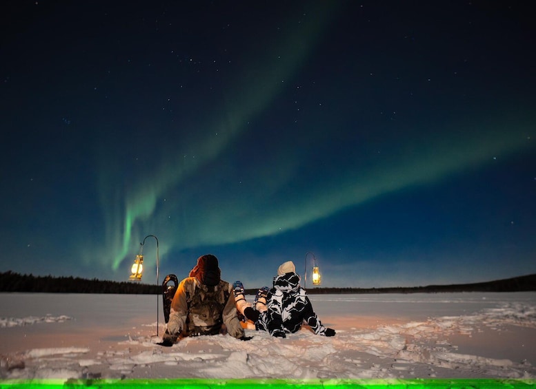 Two people sitting on a frozen lake with the Northern Lights in the background. 5