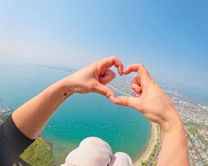A person making a heart shape with their hands, overlooking a coastal area with a clear blue sky. 1