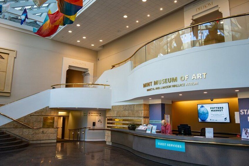 A modern interior with a reception desk labeled 'GUEST SERVICES' and a sign for the 'Mint Museum of Art.' 2