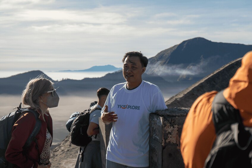 A person in a white t-shirt with 'YIXPLORER' text, standing on a rocky outcrop with a mountainous landscape in the background. 5