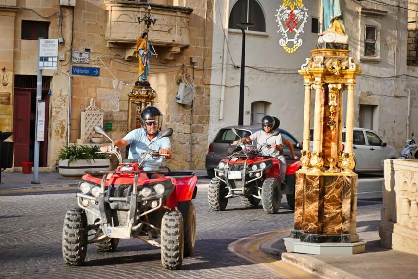 Two people riding red quad bikes on a cobblestone street, with a statue and a building in the background. 2