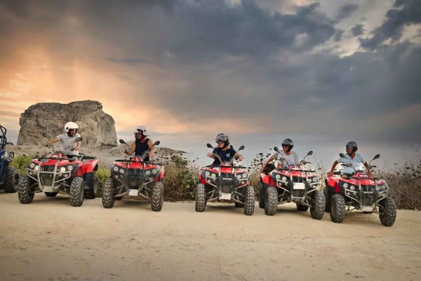 Five people on red ATVs parked on a dirt road with a rocky outcrop in the background and a cloudy sky. 3
