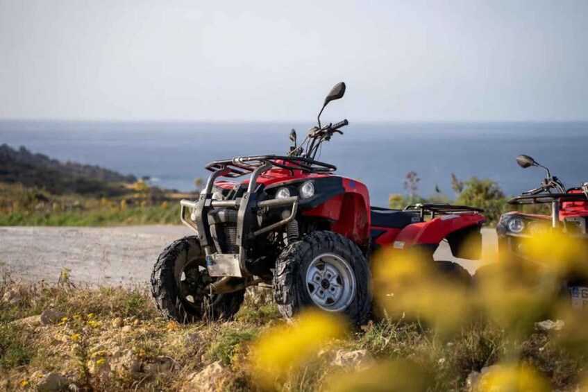 Two red ATVs parked on a dirt path with a coastal view in the background. 1