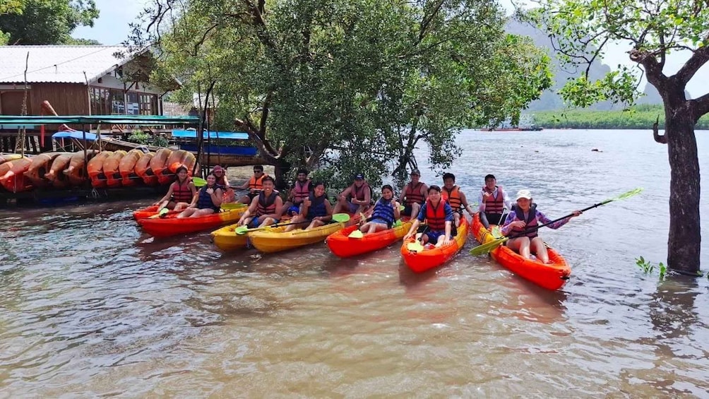 A group of people in colorful kayaks on a calm river, surrounded by trees and a dock with boats. 3