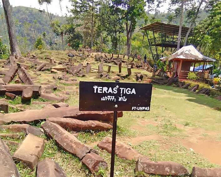 A sign reading "TERAS TIGA" in a forested area with scattered logs and a small structure in the background. 5