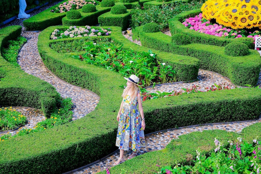 A woman in a patterned dress and hat walking on a stone pathway through a geometrically shaped garden with various flowers and hedges. 5