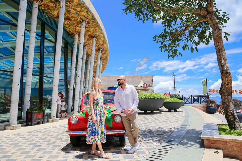A couple standing by a colorful vintage car in front of a modern building with glass windows and a curved roof. 4