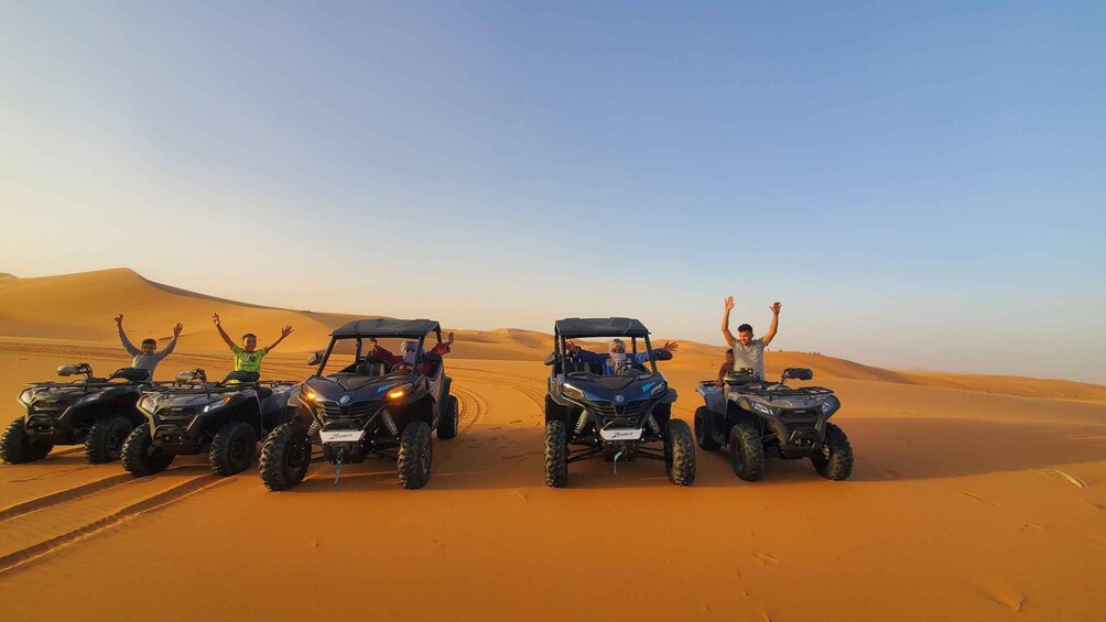 Four off-road vehicles on a desert sand dune with people raising their hands in celebration. 1
