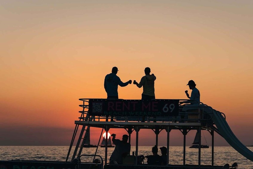 Un tramonto con le sagome di persone sul ponte di una barca, un cartello con la scritta "RENT ME 69 BEACH WEEKEND" e una barca a vela sullo sfondo. 4