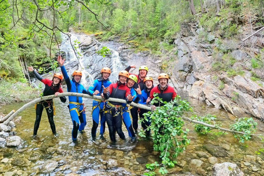 A group of people in blue and red wetsuits crossing a shallow stream over a fallen tree branch in a forested area with a waterfall in the background. 1