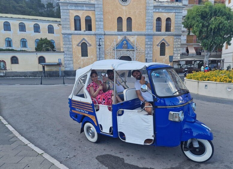A blue and white tuk-tuk with passengers, parked in front of a historic building with a bell tower. 2