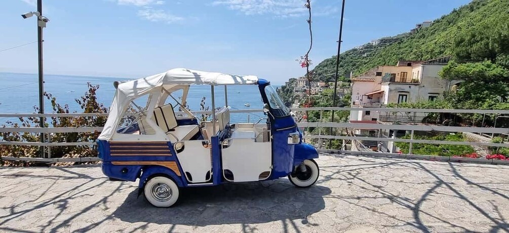 A blue and white three-wheeled vehicle parked on a paved area with a coastal view. 1