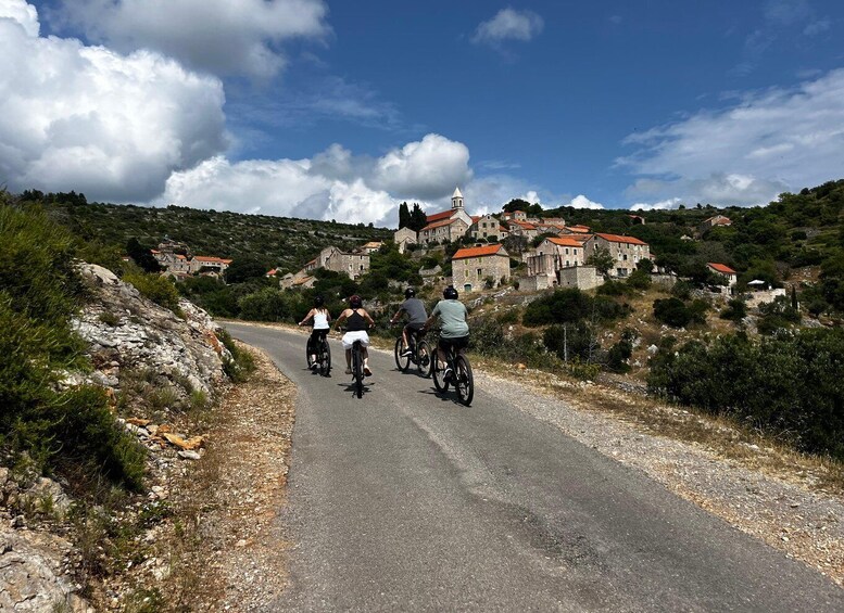 A group of cyclists on a rural road with stone houses and a church in the background. 1