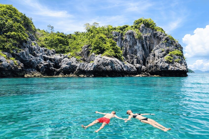 Two people floating on a turquoise sea with a rocky cliff and lush greenery in the background. 5