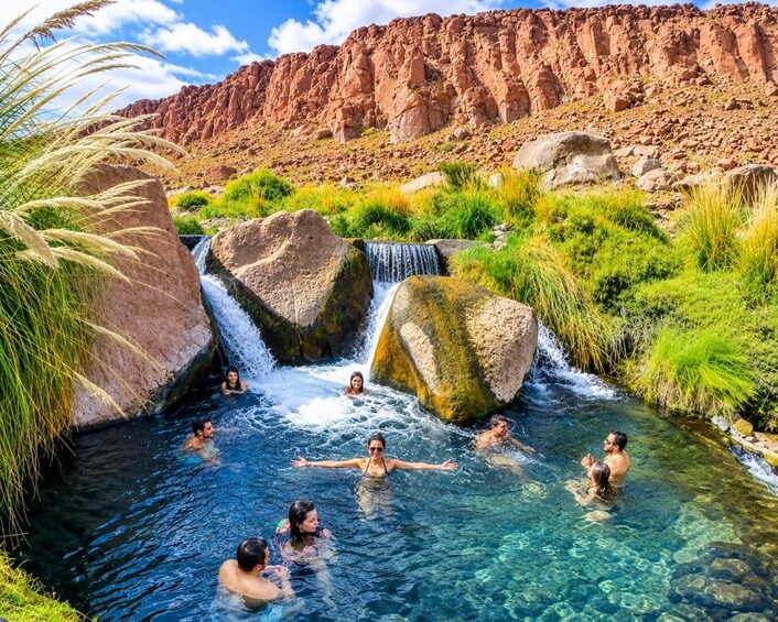 A group of people swimming in a clear, rocky stream surrounded by lush greenery and a rugged cliff. 1