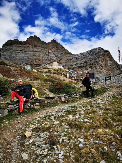 A mountainous landscape with a rocky path, a person sitting on a rock, and another person standing with a backpack. 5