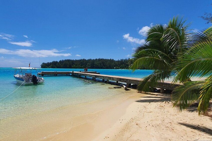 Ein Strand mit klarem türkisblauem Wasser, ein Boot an Land, ein hölzerner Steg und eine Palme. 1