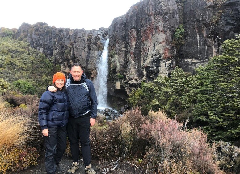 Two people standing in front of a waterfall with rocky cliffs and vegetation. 5