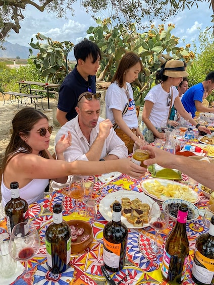 A group of people dining outdoors at a table with a colorful tablecloth, surrounded by various bottles of wine and plates of food. 5