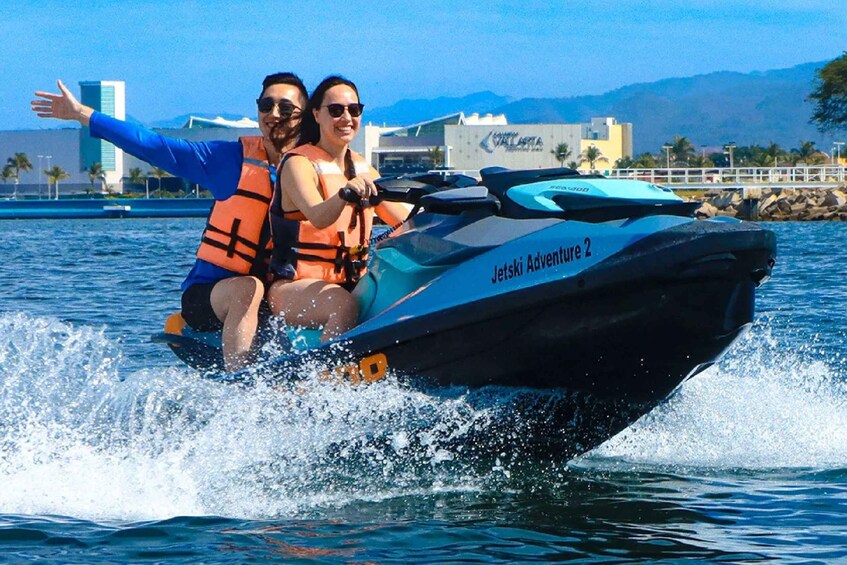 Kaksi henkilöä osallistuu Jet Ski Adventure 2 -retkeen, heillä on pelastusliivit päällään, ja taustalla näkyy vesistö sekä rakennuksia. 2