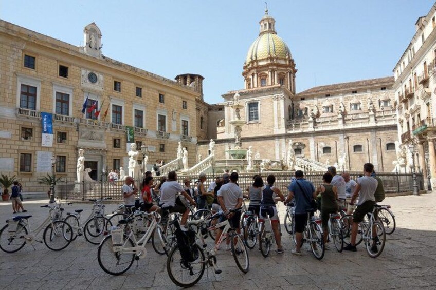 A group of people on bicycles in a plaza with historic buildings and a prominent dome in the background. 1