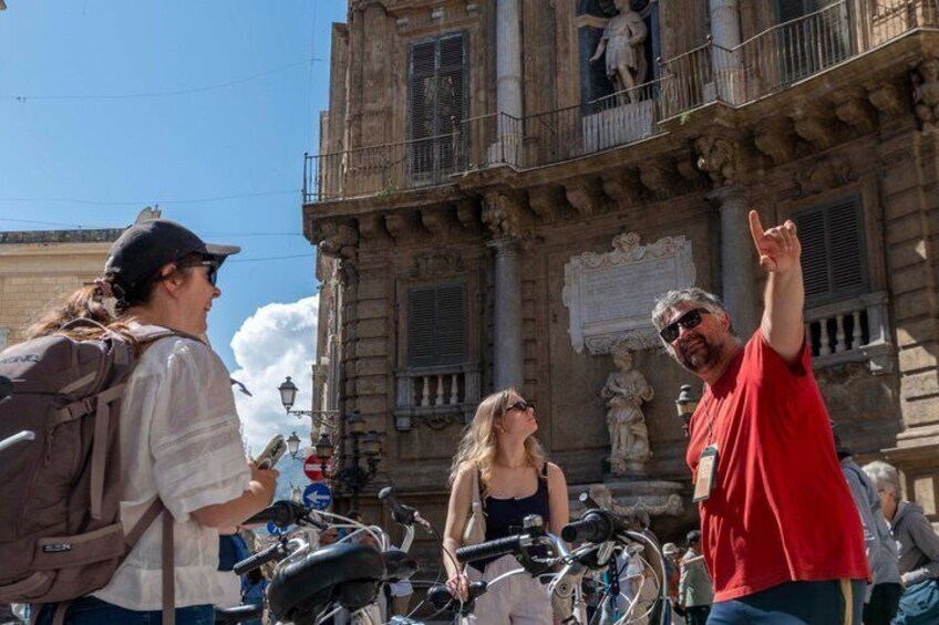 Un grupo de personas con bicicletas frente a un edificio histórico con estatuas y balcones. 1