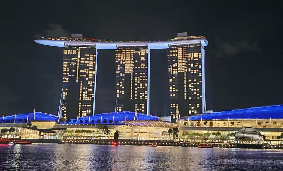 Vue nocturne d’une structure architecturale moderne, avec des fenêtres illuminées et un design de toit original, située au bord d’un plan d’eau. 1