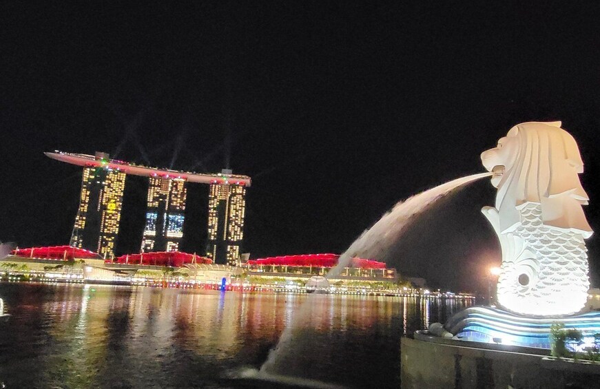 Une vue nocturne d’une ville, avec un bâtiment imposant dont les fenêtres sont illuminées, et une grande sculpture lumineuse représentant une créature marine. 2