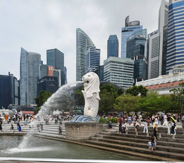 Une grande statue de lion blanc, d’où jaillit de l’eau, est entourée d’une foule de personnes, et des gratte-ciel modernes se dressent en arrière-plan. 3