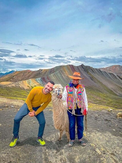 Two people with a llama on a rocky terrain with mountains in the background. 2