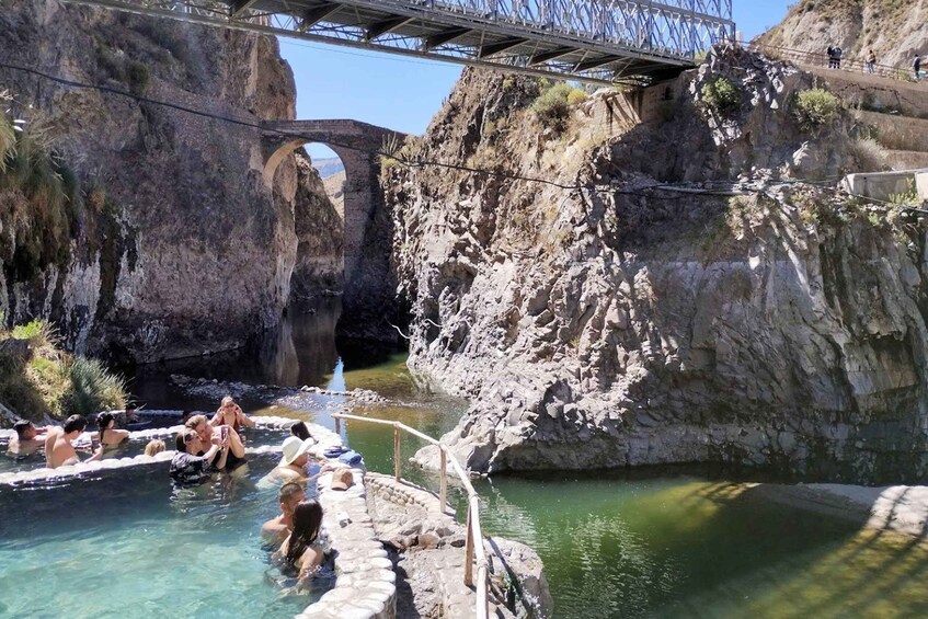A natural hot spring with people relaxing in the water, surrounded by rocky cliffs and a bridge in the background. 2