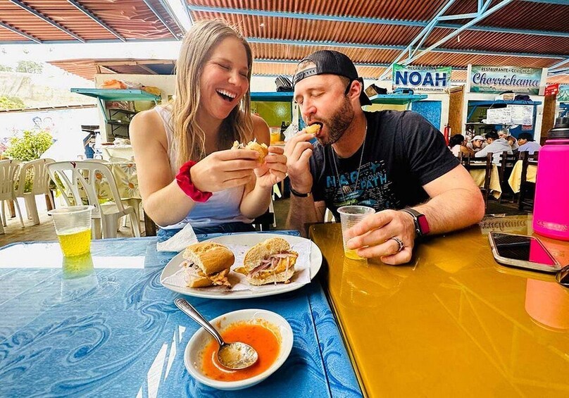 Two people eating at an outdoor restaurant with a blue tablecloth, a plate of food, a drink, and a spoon. 1