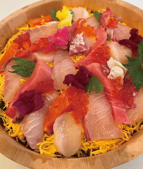 A wooden bowl containing various types of sashimi, including salmon, tuna, and octopus, garnished with flowers and shredded yellow vegetable. 3