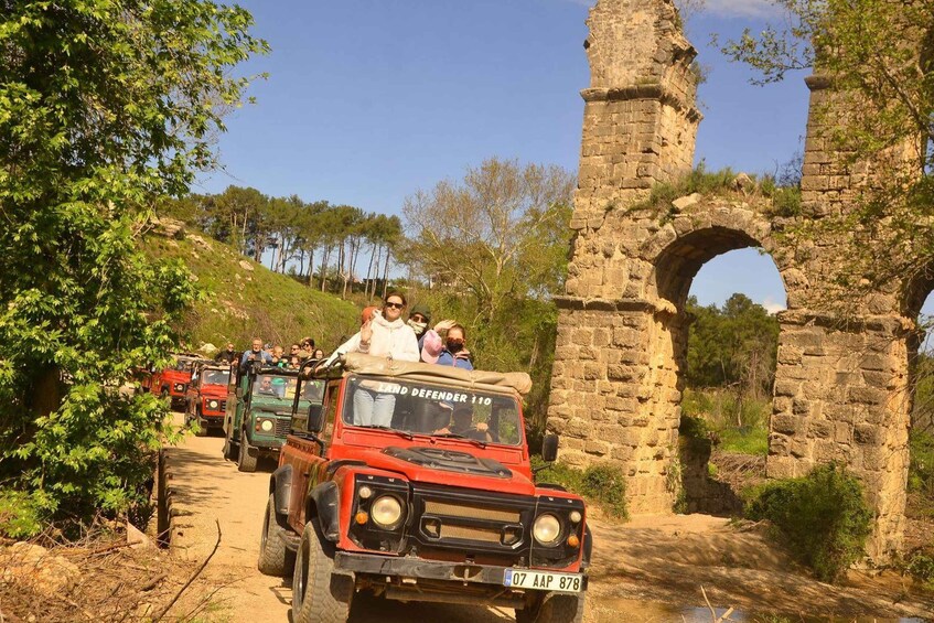 A group of tourists on a Land Defender 110 vehicle, driving past ancient stone ruins and a large archway. 2