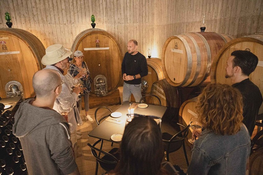 A group of people in a wine cellar with wooden barrels, a table with glasses, and a bottle of wine. 1