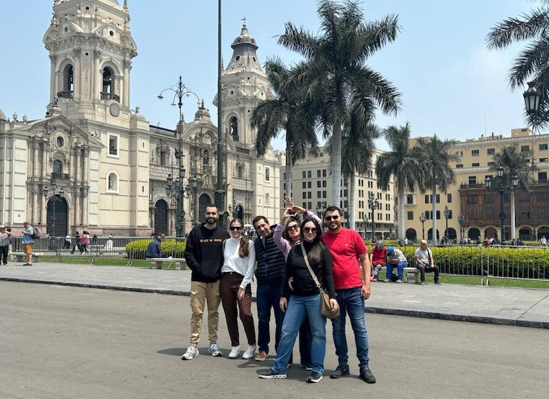 A group of people posing in front of a historic building with two towers and palm trees in the background. 1