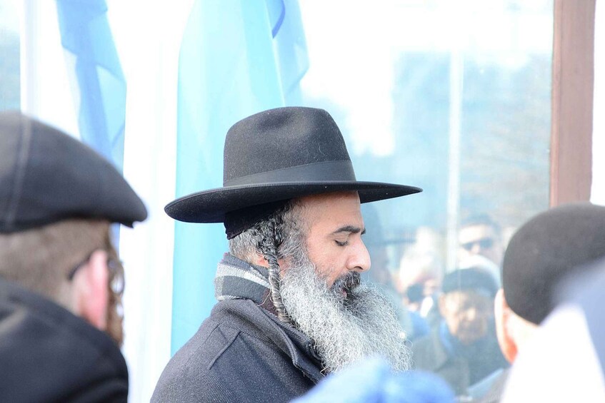 A person wearing a black hat and a long white beard, surrounded by others in similar attire. 1