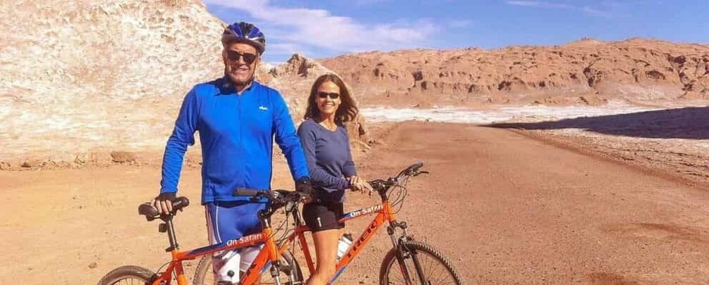Dos personas de pie junto a una bicicleta en un camino de tierra en un paisaje desértico. 4