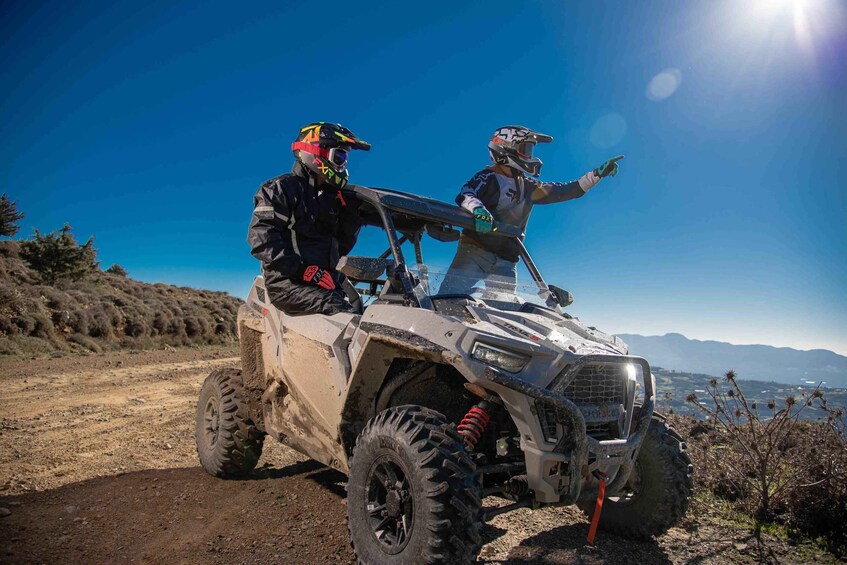 Two people on an ATV in a rugged outdoor setting with mountains in the background. 1