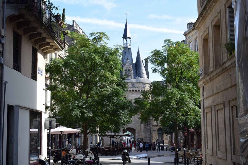 A historic street with a prominent church tower, trees lining the sidewalk, and outdoor cafes. 1